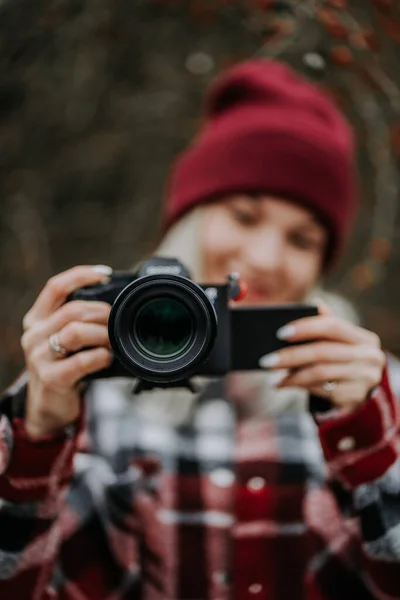 Portrait of young woman using camera in forest. Girl filming and smiling, lady in trendy shirt and red beanie hat. — Stock Photo, Image