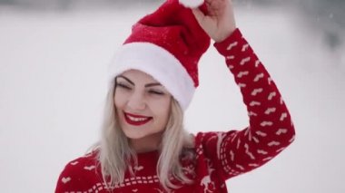 Noel süveteri ve Noel Baba şapkası içinde güzel bir kadın portresi. Kız gülümsüyor, dans ediyor, kameraya hava öpücüğü gönderiyor..
