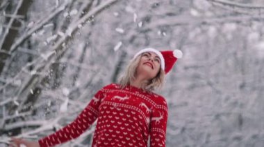 Neşeli kadın ağacın dalını çeker, üstünde kar uçar ve kışın ve soğuk havanın keyfini çıkarır. Kırmızı Noel kazağı ve Noel Baba şapkalı kız. Yavaş çekim.