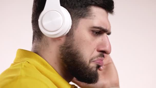 Hombre guapo con barba con auriculares se pone alto en la música, se mueve al ritmo, canta junto con la canción en el estudio sobre fondo blanco. Danza activa, concepto de radio, cámara lenta — Vídeo de stock