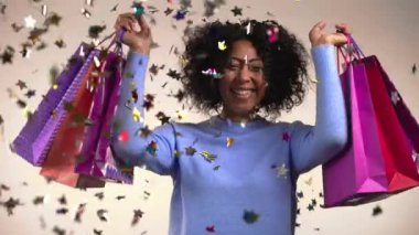 Stüdyo arka planında konfeti yağmurunun üzerinden atladıktan sonra renkli kese kağıtlarıyla heyecanlı Afrikalı kadın. Mevsimlik satış kavramı, alışverişler, hediyelere para harcamak.