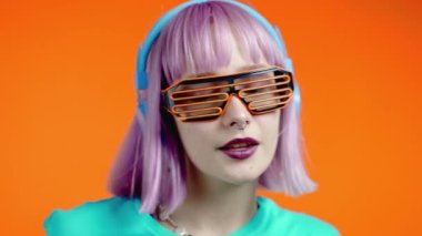 Alışılmadık moda bir kadın, boyalı pembe saçlı müzik dinliyor, turuncu arka planda kulaklıkla dans ediyor. Renk engelleyici. Kafes gözlüklü modacı kız. Yavaş çekim.