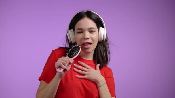 Jovem mulher bonita cantando e dançando com escova de cabelo ou pentear em vez microfone na parede violeta. Menina em fones de ouvido se divertindo, ouvindo música, sonhos de ser celebridade. — Vídeo de Stock