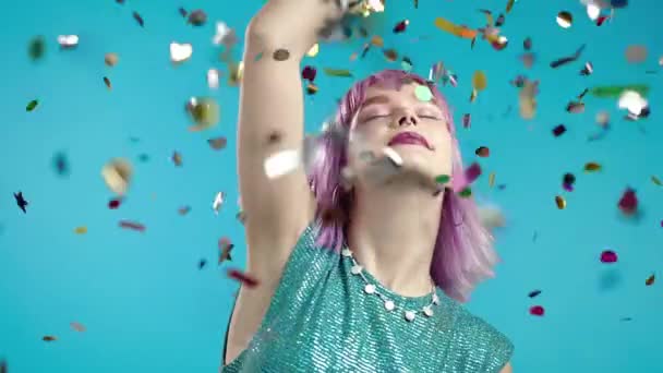 Mulher moderna alegre dançando, tocando o cabelo tingido violeta incomum durante a chuva de confetes no estúdio azul. Conceito de comemorar, festa, ganhar — Vídeo de Stock