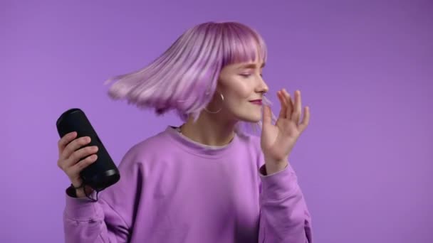 現代の流行の女の子は、ワイヤレスポータブルスピーカーで音楽を聴く。紫の髪型の若い美しい女性がスタジオの背景で楽しんで踊っています。女性は音楽のリズムに移行します. — ストック動画