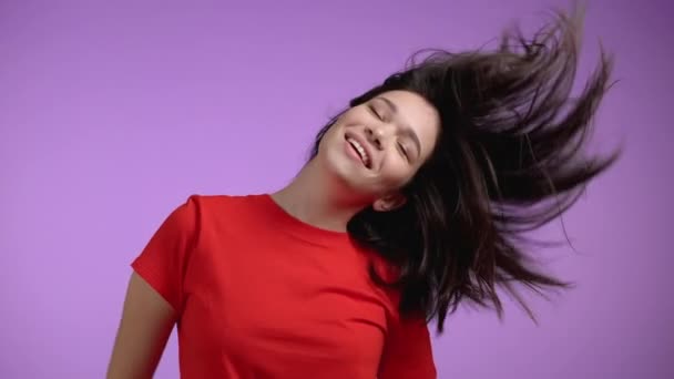 Cute kobieta taniec z głową na fioletowym tle studio, jej włosy trzepotanie.Dziewczyna w pomarańczowym t-shirt. Pozytywny nastrój. Zwolniony ruch. — Wideo stockowe