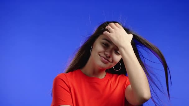Aantrekkelijke vrouw poseren op blauwe studio achtergrond, haar lange haren fladderen uit de luchtstroom. Meisje in kleurrijke oranje kleding. Positieve stemming. Langzame beweging. — Stockvideo