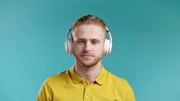 Beau jeune homme écoutant de la musique avec des écouteurs sans fil, gars qui s'amuse, souriant en studio sur fond bleu. Danse, concept radio. — Video