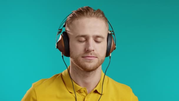 Homem loiro ouvindo música com fones de ouvido, cara se divertindo, sorrindo em estúdio em fundo azul. Dança, conceito de rádio. — Vídeo de Stock