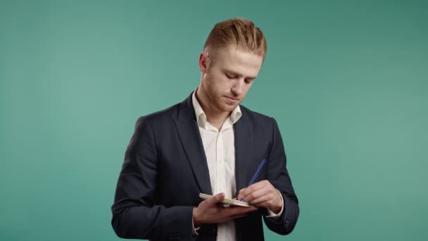 Left-handed young businessman making notes in planner, guy holding pen, reflects on plans. Professional person in suit makes new appointments with clients on blue studio background. — Stock Video