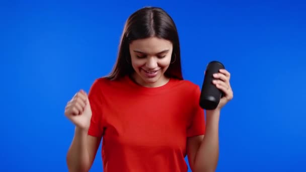 Vrouw die luistert naar muziek door draadloze draagbare luidspreker - modern geluidssysteem. Jong meisje dansend, genietend op een blauwe studio achtergrond. Ze beweegt naar het ritme van de muziek. — Stockvideo