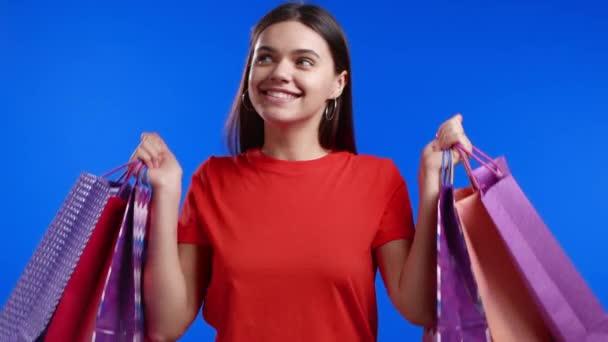 Excited woman with colorful paper bags after shopping on blue studio background. Concept of seasonal sale, purchases, spending money on gifts — Stock Video