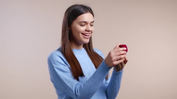 Attractive woman holding small jewelry box with proposal diamond ring on light wall background. Lady smiling, she is happy to get present, proposition for marriage — Stock Video