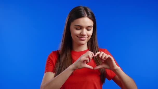 Mulher bonita gentil fazendo sinal de forma coração perto de seu peito. Fundo azul brilhante. Saúde da mulher, voluntariado, ajuda de doação e conceito de amor. — Vídeo de Stock