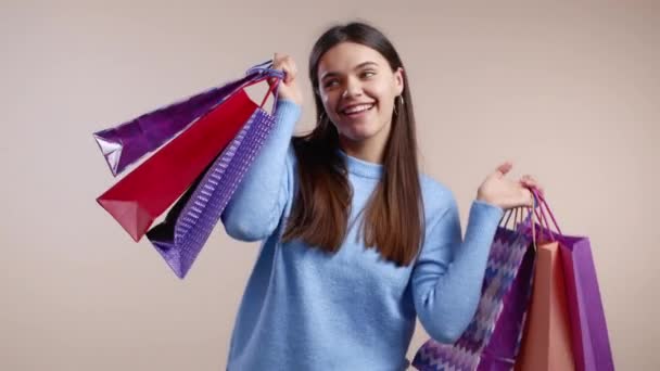 Renkli kağıt torbalarla bej renkli stüdyo arka planından alışveriş yapan heyecanlı bir kadın. Mevsimlik satış kavramı, alışverişler, hediyelere para harcamak. — Stok video