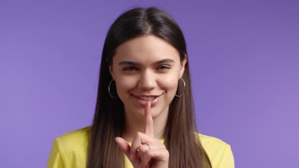Жест, таємниця, тиша. Усміхнена жінка тримає палець на губах над фіолетовим тлом. крупним планом . — стокове відео