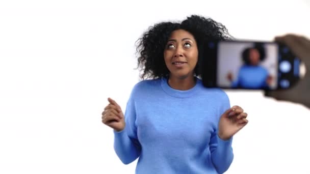 Uśmiechnięty blogerka afrykańska kobieta tworząc online nowy blog wideo, vlog, ze swoim smartfonem na białym tle. Influencer przemawiający do kamery podczas nagrywania rozmów konferencyjnych. — Wideo stockowe