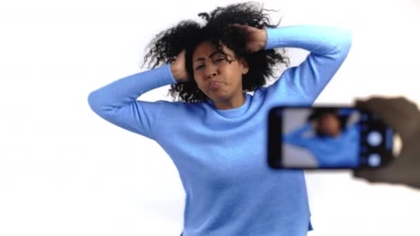 Divertida vlogger mujer con el pelo afro grabación de vídeo de sí misma bailando delante de la cámara del teléfono inteligente sobre fondo blanco. Influenciador de raza mixta hace clip de redes sociales — Vídeo de stock