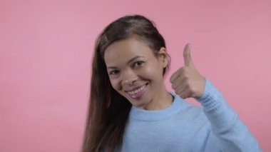 Afrikalı kadın pembe arka planda baş parmak işareti gösteriyor. Olumlu genç kız kameraya gülümsüyor. Kazandım. Başarı. Vücut dili.