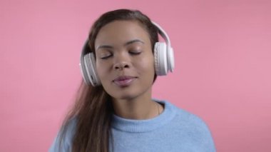 Pembe stüdyo arka planında izole edilmiş kablosuz kulaklıklarla dans eden melez bir kadın. Hoş bir bayan portresi. Müzik, radyo, mutluluk, özgürlük, gençlik kavramı.