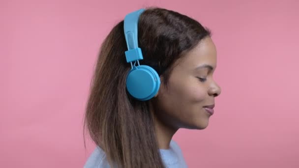 Vegyes rasszista nő táncol vezeték nélküli kék fejhallgató elszigetelt rózsaszín stúdió háttér. Aranyos női portré. Zene, rádió, boldogság, szabadság, ifjúságpolitika. — Stock videók