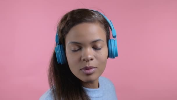 Blandad ras kvinna dans med trådlös blå hörlurar isolerad på rosa studio bakgrund. Gulligt kvinnoporträtt. Musik, radio, lycka, frihet, ungdomsidé. — Stockvideo
