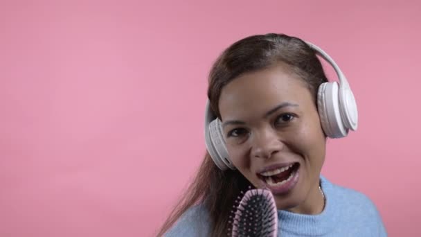 Afrikanische Frau singt und tanzt mit Haarbürste oder Kamm statt Mikrofon an rosa Wand. Dame mit Kopfhörer hat Spaß, hört Musik, träumt davon, berühmt zu werden. — Stockvideo