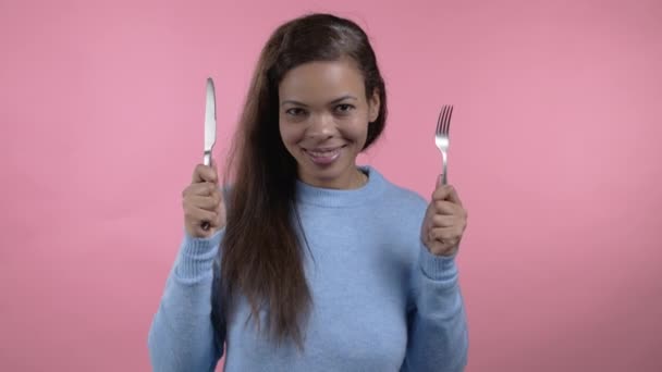 Portret głodnej Afrykanki z widelcem i nożem. Dziewczyna czeka na serwowanie dań obiadowych z sztućcami na różowym tle studio. — Wideo stockowe