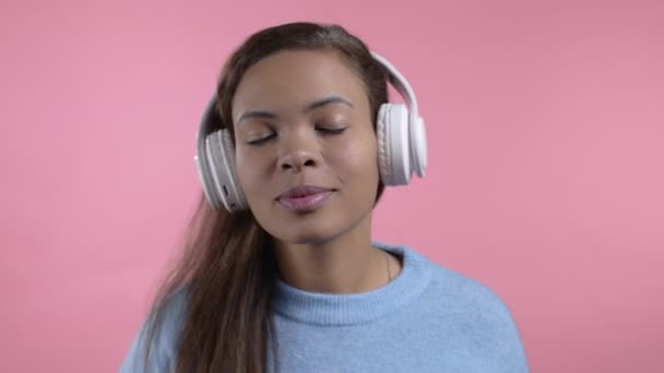Gemengde race vrouw dansen met draadloze hoofdtelefoon geïsoleerd op roze studio achtergrond. Schattig vrouwenportret. Muziek, radio, geluk, vrijheid, jeugdconcept. — Stockvideo