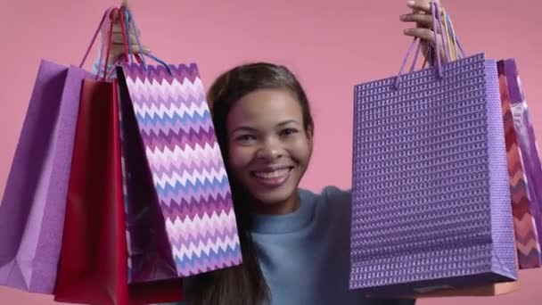 Femme africaine excitée avec des sacs en papier colorés après avoir fait ses courses sur fond de studio rose. Concept de vente saisonnière, achats, dépenses en cadeaux — Video