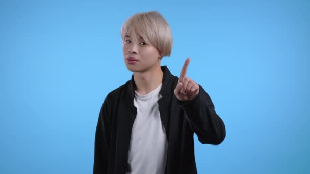 Egy komoly koreai férfi portréja, amint elutasító gesztust mutat a mutatóujjával. Ázsiai srác festett szőke haj elszigetelt kék háttér. — Stock videók