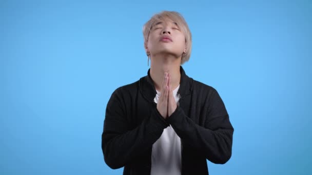Fiatal ázsiai férfi, kék háttérrel imádkozik. Koreai stílusban könyörög valakinek, hogy elégítse ki a vágyait, segítsen.. — Stock videók