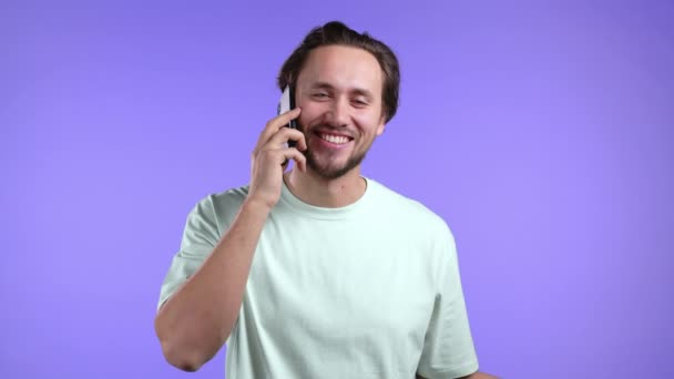 El joven habla con una sonrisa por teléfono. Un tipo sosteniendo y usando un teléfono inteligente. Estudio violeta fondo. — Vídeo de stock
