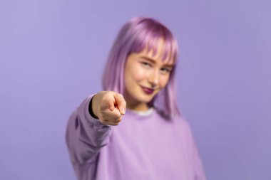 Mor renkli saçları olan çekici bir kadın işaret parmağıyla kamera lensine işaret ediyor ve göz kırpıyor, flört ediyor. Hey, sen menekşe arka plandaki kız..