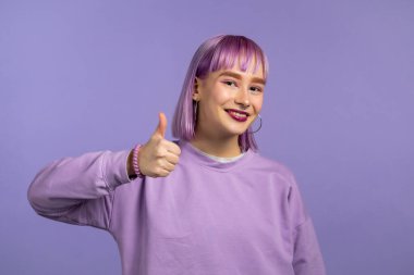 Modaya uygun bir kadın, Violet stüdyosunun arka planında işaret gibi görünüyor. Mor saçlı pozitif genç kız kameraya gülümsüyor. Kazandım. Başarı. Vücut dili.