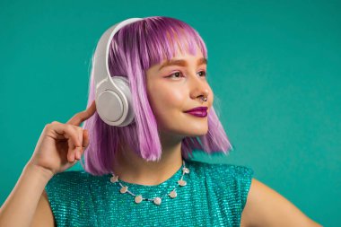 Alışılmadık modaya uygun bir kadın menekşe rengi saçlarıyla müzik dinliyor, turkuaz arka planda kulaklıkla dans ediyor. Moda tutkunu genç kız.