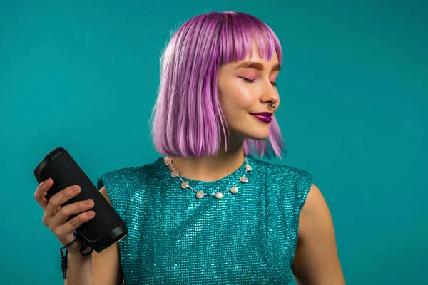 Unusual woman with dyed violet hairstyle listening to music by wireless portable speaker - modern sound system. Young girl dancing, enjoying on turquoise studio background