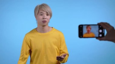 Gülümseyen Koreli blogcu adam çevrimiçi yeni bir video blogu yapıyor, mavi arka planda akıllı telefonuyla video blogu yapıyor. Konferans kayıtları sırasında kameraya konuşan nüfuzlu kişi.