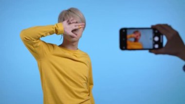Gülümseyen Asyalı vlogger adam mavi arka planda akıllı telefon kamerasının önünde dans ederken videosunu kaydediyor. Nüfuzlu biri komik sosyal medya klipleri yapıyor. K-pop kültürü.