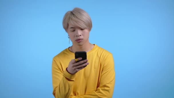 Jóképű koreai férfi okostelefont használ a kék stúdió falán. Szőke fickó sárga ruhában, modern technológiával - alkalmazások, közösségi hálózatok. — Stock videók