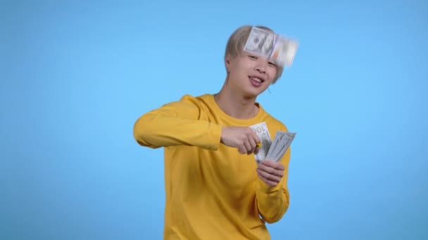 Uomo asiatico con la faccia felice disperde i soldi. Un coreano spende troppo in dollari. E 'pieno di dollari su sfondo blu. — Video Stock