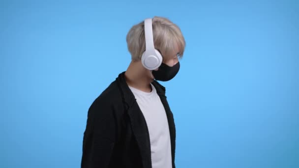 Νεαρός με μάσκα και μοδάτο χτένισμα χορεύει με ακουστικά στο στούντιο με φόντο το μπλε. Μουσική, χορός, ραδιοφωνική έννοια. — Αρχείο Βίντεο