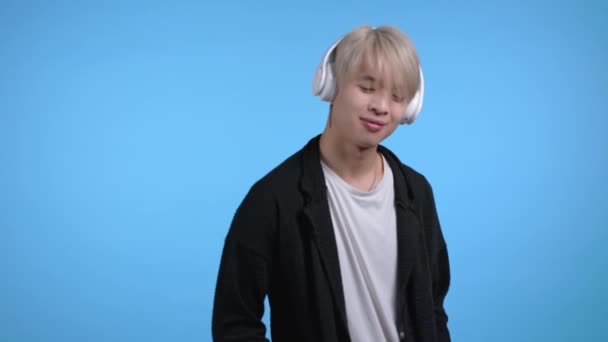 Hombre coreano guapo con peinado rubio de moda divirtiéndose, sonriendo con auriculares en el estudio sobre fondo azul. Música, danza, concepto de radio. — Vídeo de stock