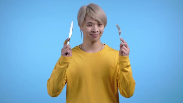 Portret van hongerige Aziaat met vork en mes. Kerel wachtend op het serveren van servies met bestek op blauwe studio achtergrond. — Stockvideo