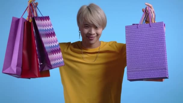 Schöner asiatischer Mann hält Einkaufstüten auf blauem Studiohintergrund. Kerl kaufte Geschenke mit Rabatten. — Stockvideo