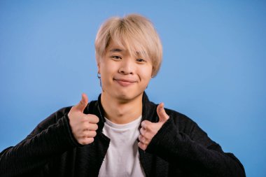 Yakışıklı Koreli adam baş parmağını kaldırıyor, jest gibi. Mavi arka plandaki mutlu Asyalı adam. Kazandım. Başarı. Pozitif erkek model kameraya gülümser. Vücut dili.