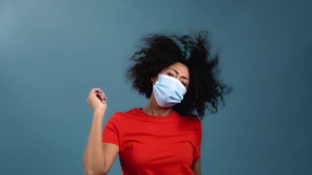 Gemengde race vrouw met afro kapsel dansen in gezichtsmasker op blauwe studio achtergrond. Mooi vrouwelijk model in oranje t-shirt. Feest, geluk, vrijheid, jeugdconcept. — Stockvideo