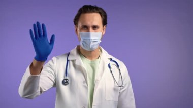 Dost canlısı doktor, lateks eldivenli el sallıyor. Merhaba. Selamlar, kameraya merhaba deyin. Violet stüdyosundaki maskeli ve ceketli yakışıklı doktor. Tıp konsepti.