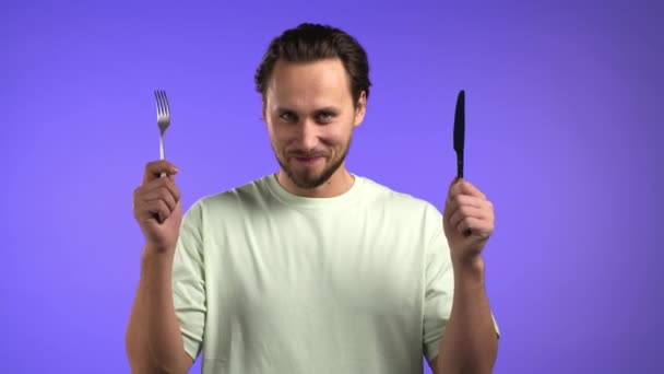 Portret głodnego człowieka z widelcem i nożem. Facet czeka na serwowanie dań ze sztućcami na fioletowym tle studia. — Wideo stockowe