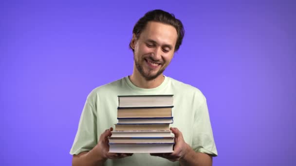 Estudiante masculino tiene pila de libros universitarios de la biblioteca sobre fondo violeta en el estudio. Guy sonríe, está feliz de graduarse. — Vídeos de Stock